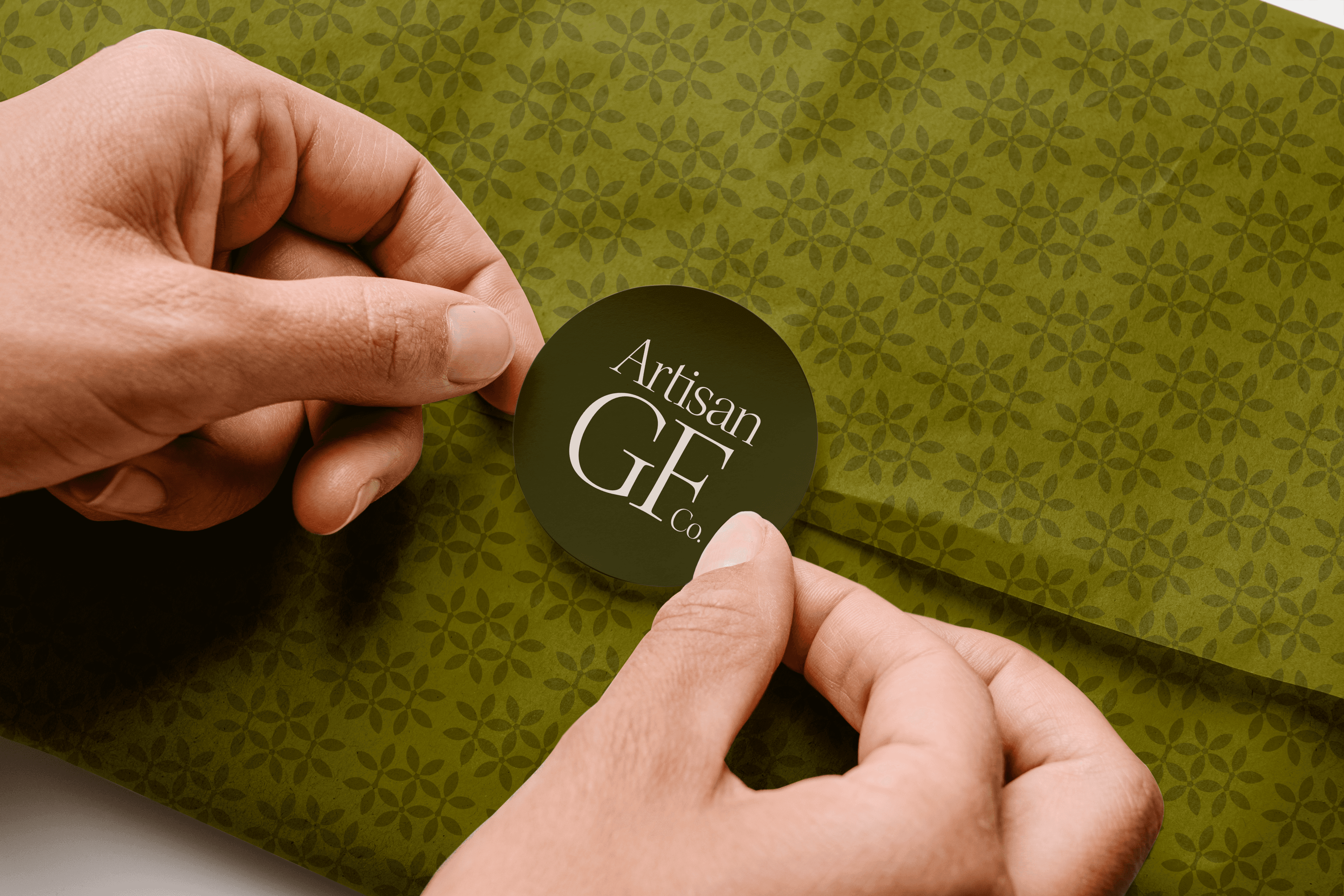 Hands pressing a round Artisan GF Co. sticker with the oversized GF lockup onto olive patterned tissue paper
